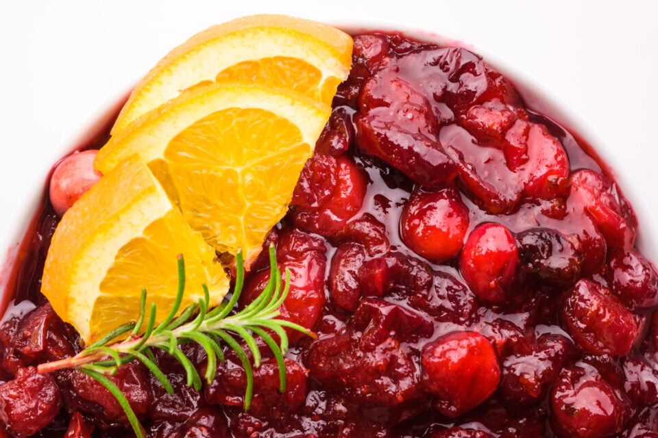Looking down on a bowl of bright red sauce with orange slices and a twig of rosemary.