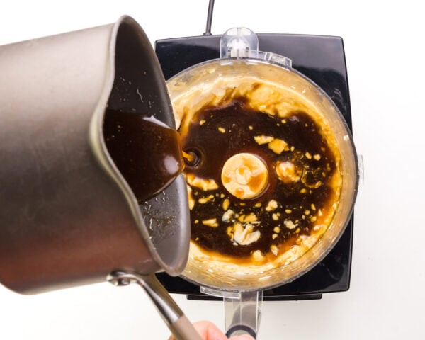 Syrup from a saucepan is being poured into a food processor with silken tofu.