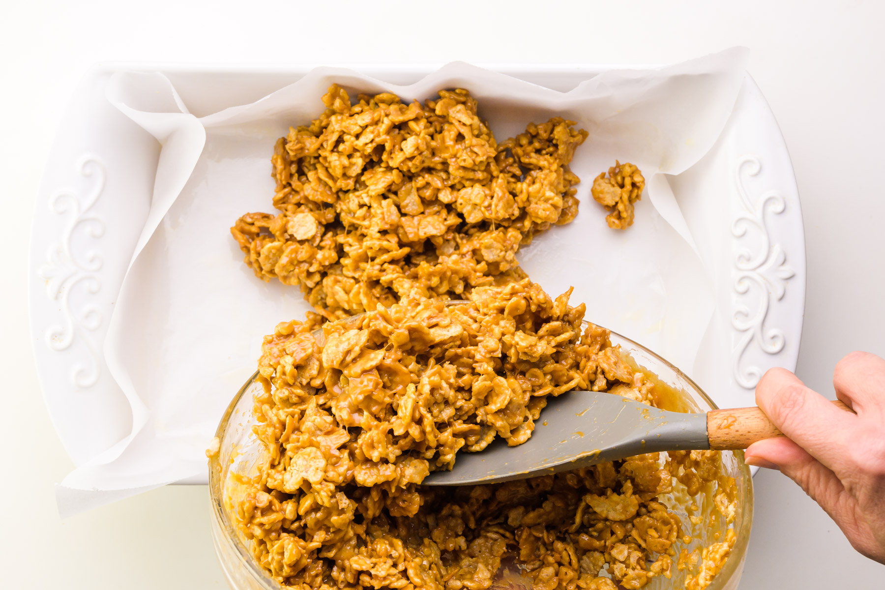 A hand holds a spatula, pressing cereal bar batter into a prepared pan.