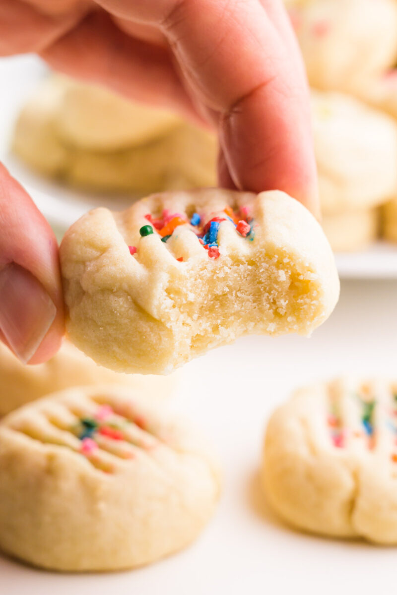 A hand holds a vegan shortbread cookie with a bite taken out. There are more cookies in the background.