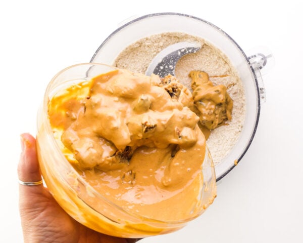 A peanut butter mixture is being poured into a food processor with oat flour.