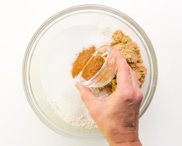 A hand pours a bowl of spices into a mixing bowl with four, sugar, and other ingredients.