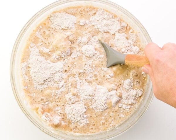 A hand holds a spatula, stirring cake batter in a bowl.