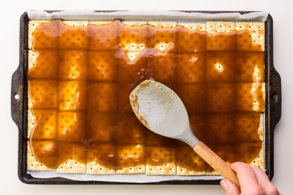 A hand holds a spatula, spreading caramel over crackers.