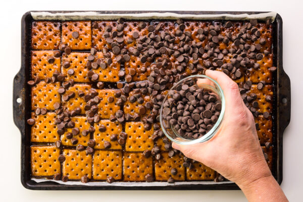 Chocolate chips are being spread over baked crackers in a pan.