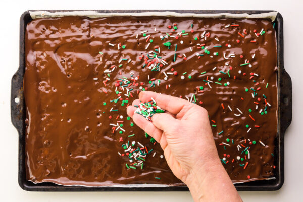 A hand spreads Christmas sprinkles over a pan with chocolate over crackers.
