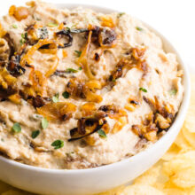 A bowl of vegan French onion dip sits next to a plate of chips.