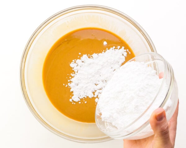 A hand holds a bowl pouring powdered sugar into a bowl with melted peanut butter.