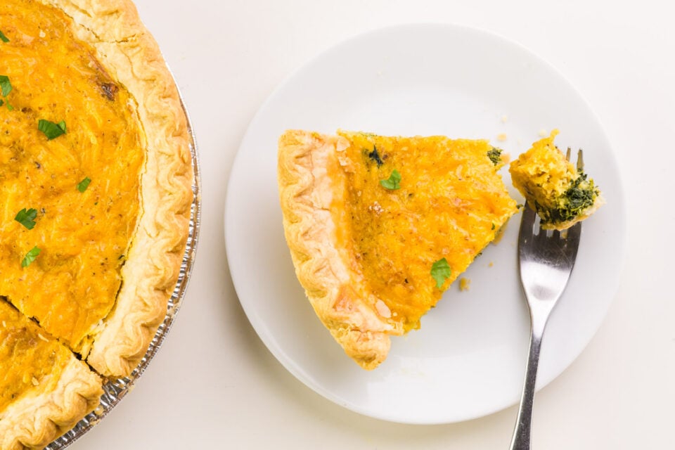 A slice of vegan quiche has a bite taken out and sits on a plate next to the rest of the quiche in the pie pan.
