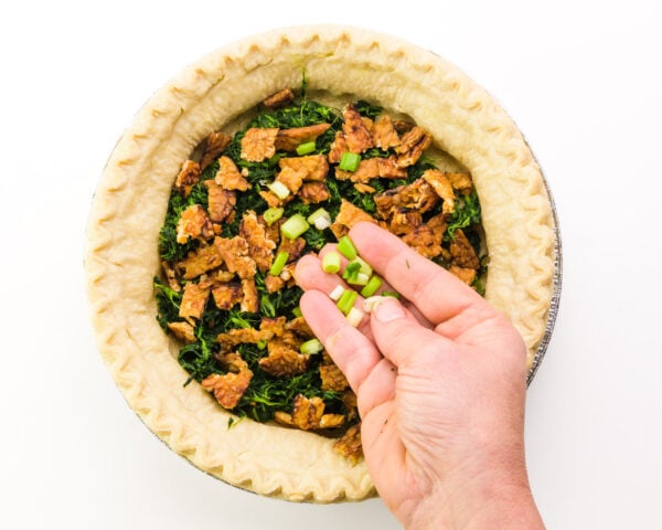 A hand spreads toppings over a pre-baked pie crust.