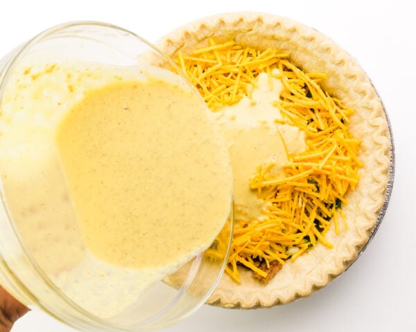 A vegan egg mixture is being poured over a pie crust with cheese and other ingredients.
