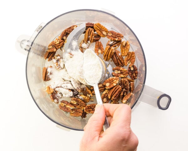 A hand holds a spoon, sprinkling flour into a food processor with toasted pecans.