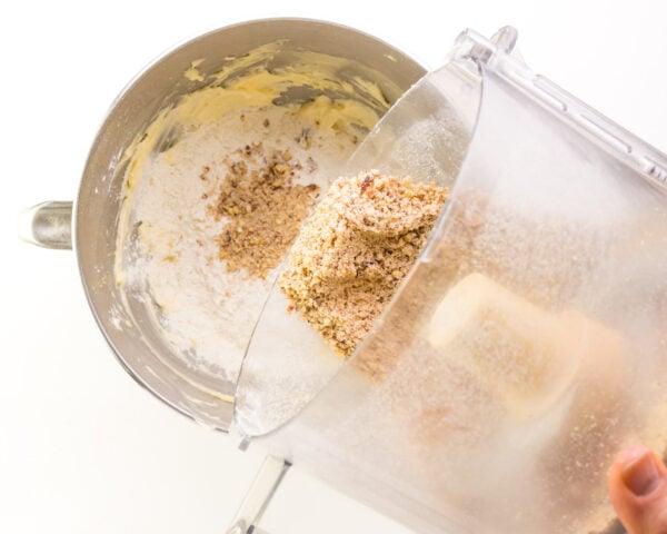 Processed nuts are being poured from a food processor bowl into a mixing bowl with cookie dough ingredients.