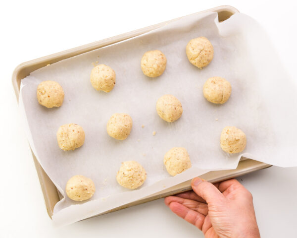 A hand holds a pan lined with parchment paper with cookie dough balls.