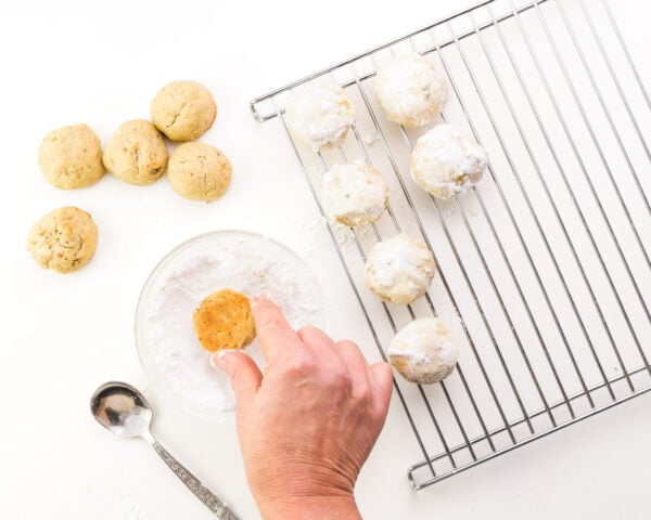 A hand rolls cookies in a bowl with powdered sugar. There are cookies on a wire rack.