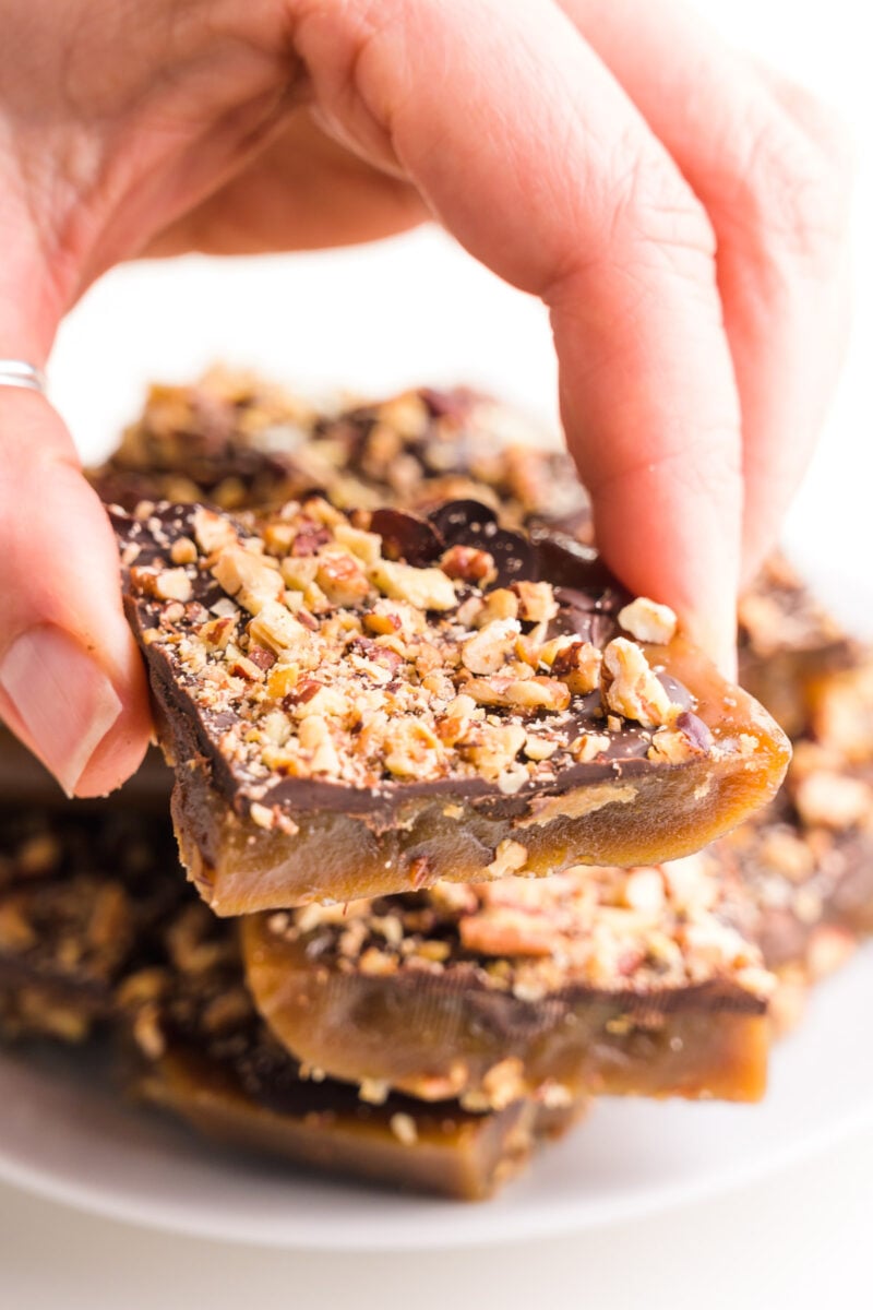 A hand holds a vegan toffee bar over a plate with more of the candy.