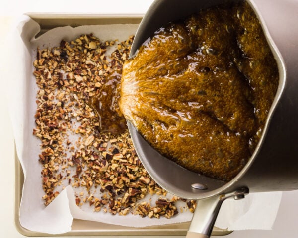 Hot sugar mixture is being poured over a pan lined with chopped nuts.