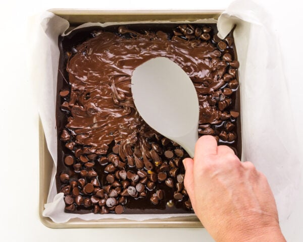 A hand holds a spatula, spreading melting chocolate chips in a pan.