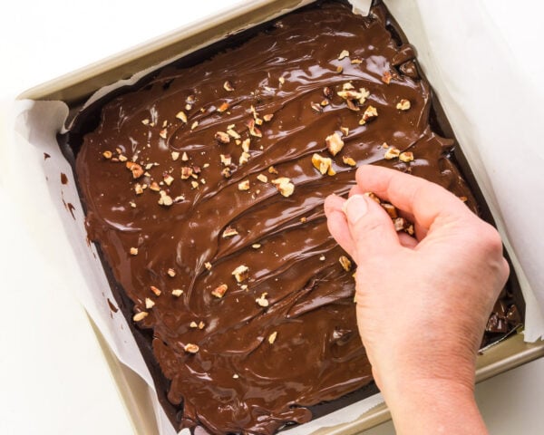 A hand spreads chopped nuts over gooey chocolate in a pan.