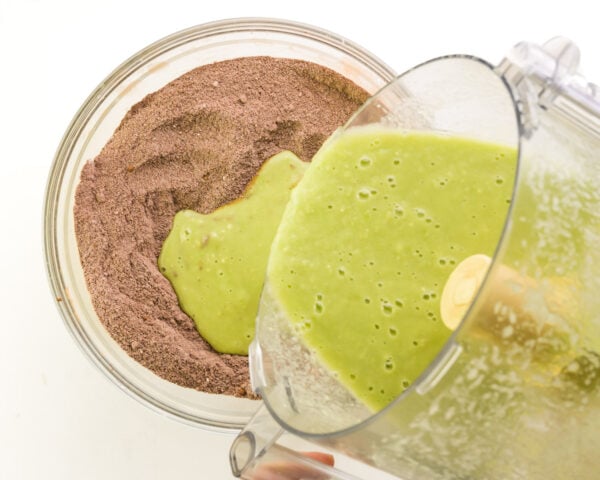 A green mixture from a food processor is being poured into a bowl with chocolate flour mixture.