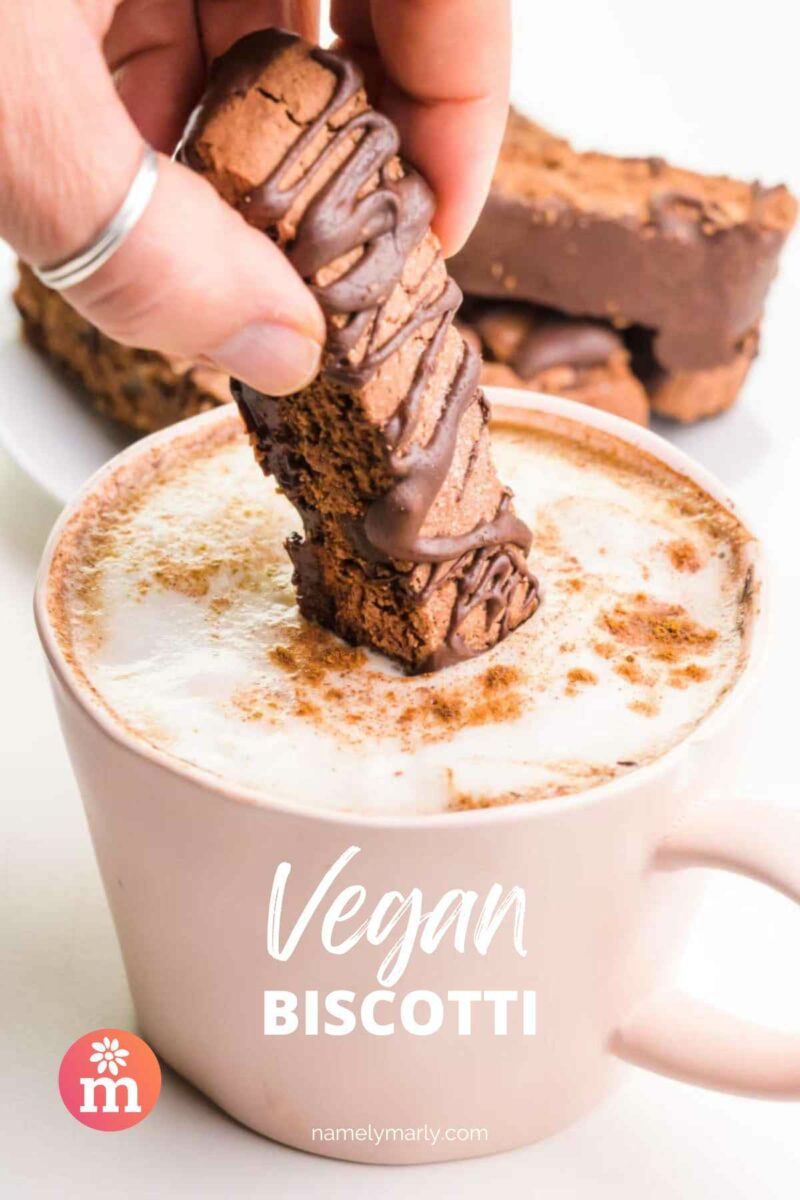 A hand dips a crisp chocolate cookie in hot tea. The text reads, Vegan Biscotti.