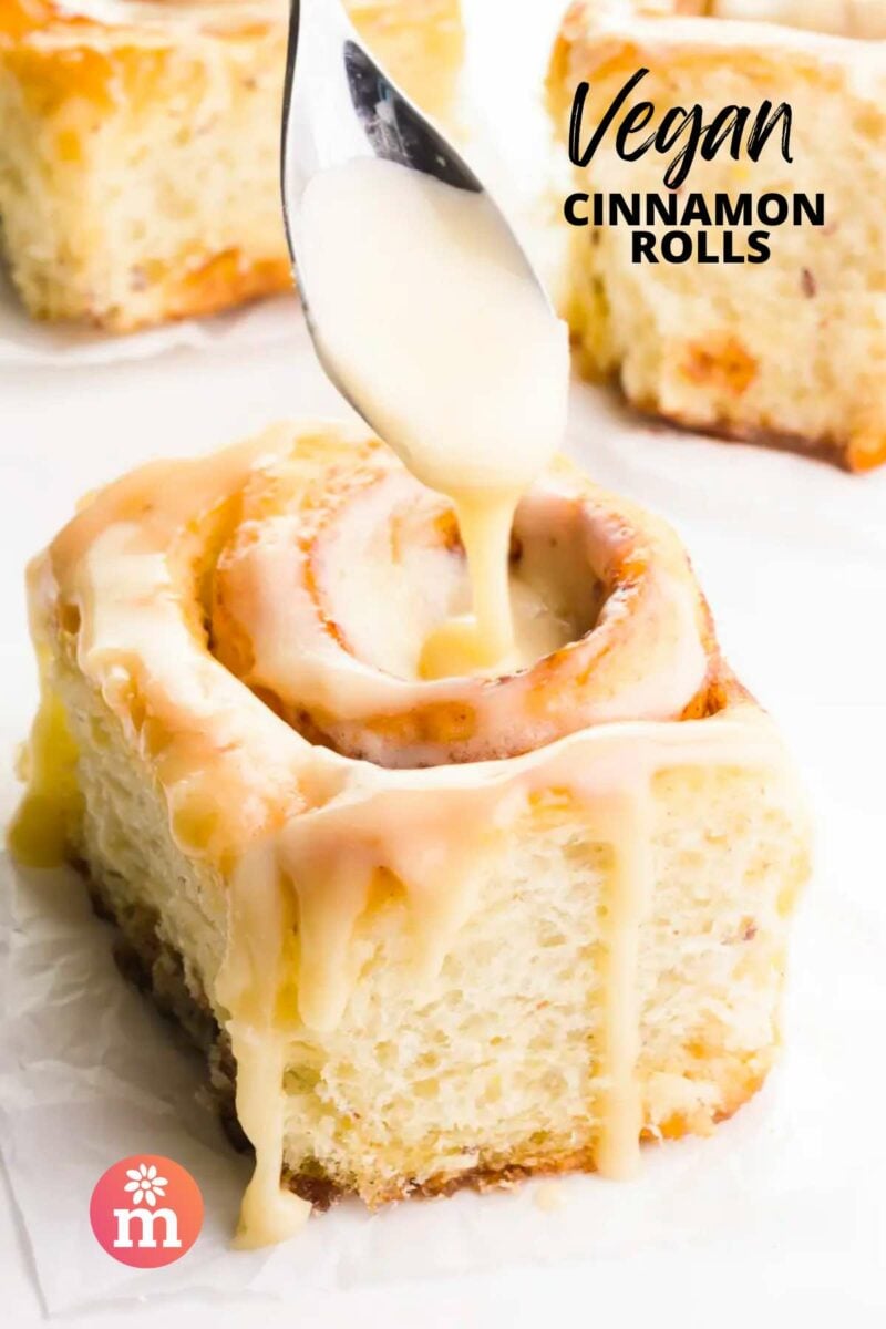 Vegan cinnamon rolls topped with a creamy glaze, with one roll drizzled from a spoon.