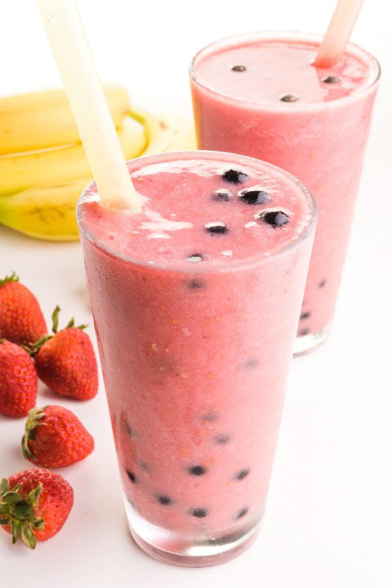 A boba smoothie is in a glass with another one behind it and fresh fruit around it.