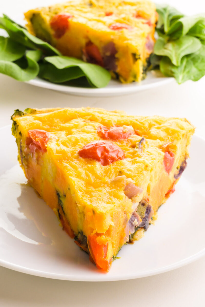 A slice of Just Egg frittata sits on a plate in front of another slice on a plate surrounded by salad greens.