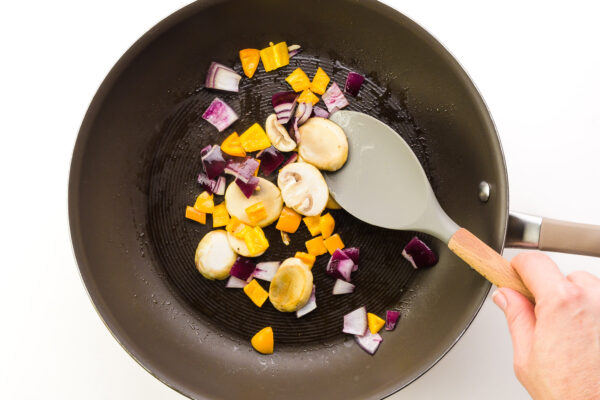 A hand holds a spatula, stirring vegetables cooking in a skillet.