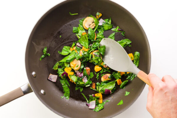 A hand holds a spatula, stirring veggies and spinach in a skillet.
