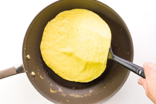 A hand uses a spatula to flip an omelet in a skillet.