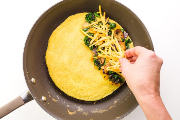 A hand distributes cheddar shreds over vegetables on one half of a whole omelet.