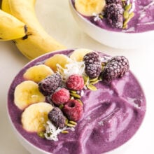 Two purple smoothie bowls have fruit on top. There are two bananas beside the bowls