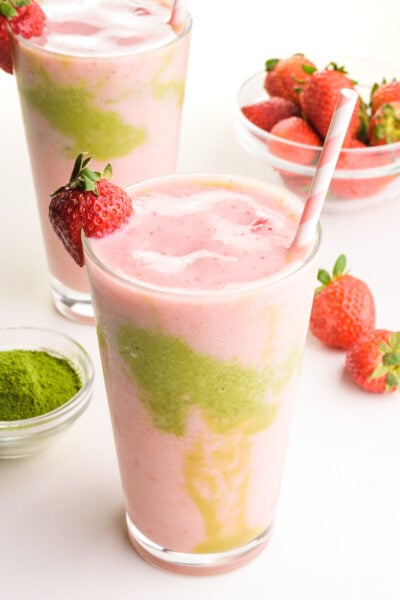 Pink and green smoothie liquid is in two glasses, one in front of the other. There is a bowl of green tea powder and strawberries around the glasses.