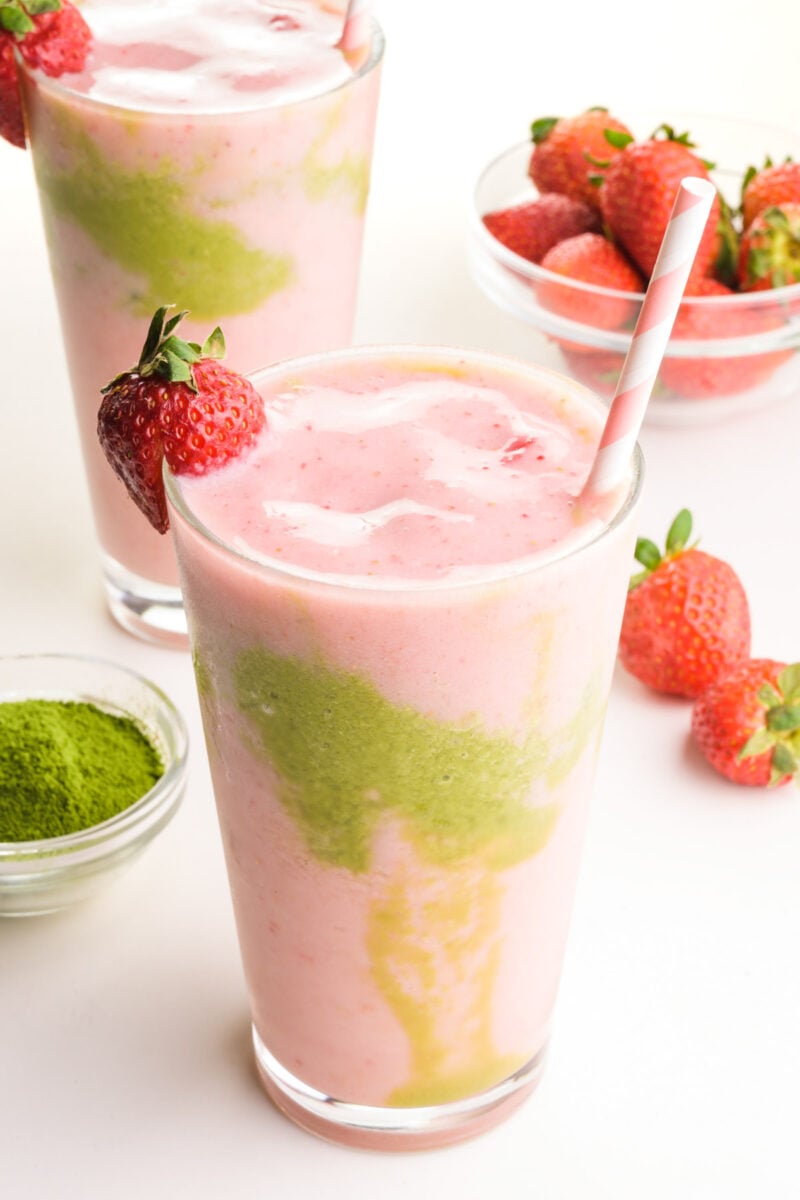 Pink and green smoothie liquid is in two glasses, one in front of the other. There is a bowl of green tea powder and strawberries around the glasses.