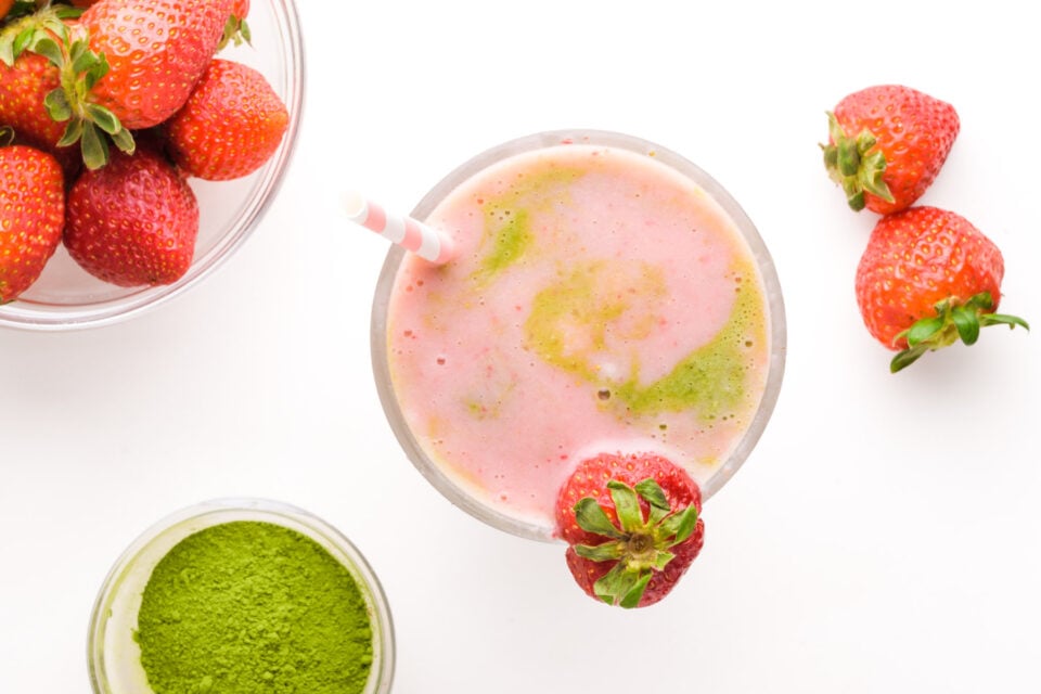 Looking down on a strawberry matcha smoothie in a glass. There are fresh strawberries and matcha powder around it.