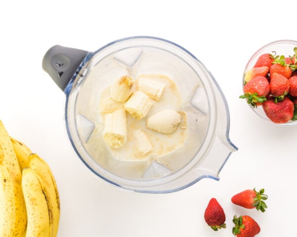 Looking down on a blender with ingredients like banana chunks. There are fresh strawberries and bananas around the blender.