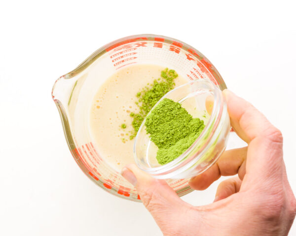 A hand pours matcha powder into a measuring cup with smoothie mixture.