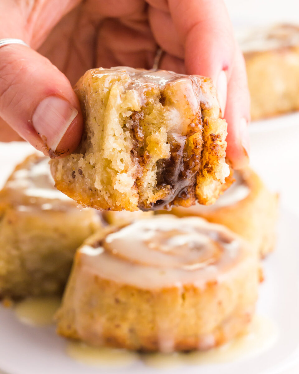 A hand holds a cinnamon roll with a bite taken out, hovering over more cinnamon rolls.