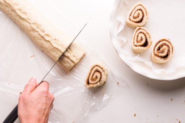 A hand holds a knife, cutting a long log of dough into slices. Some slices have been placed in a baking dish nearby.