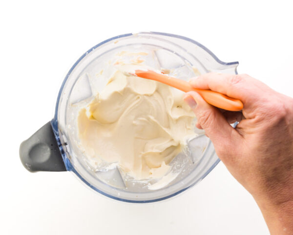 A hand holds a spatula, scraping down the sides of a blender jar with a creamy white mixture in it.