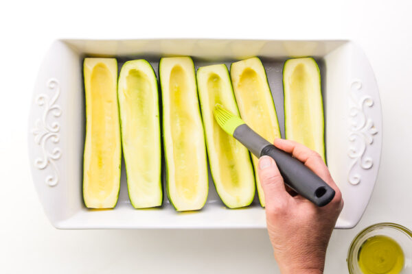 A hand holds a melon scooper, scooping flesh out of zucchinis in a baking dish.