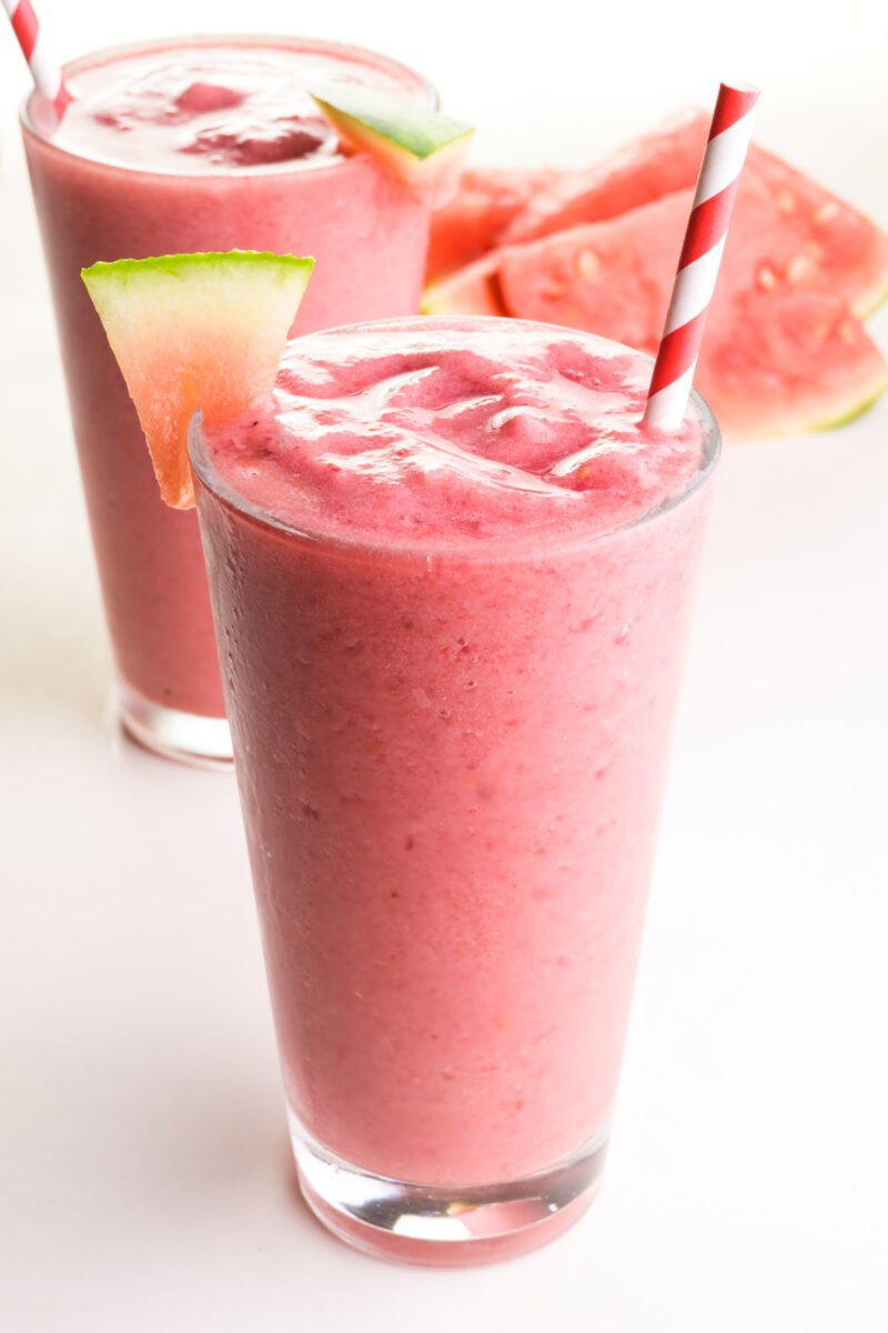 Two pink smoothies sit on a table. They have watermelon wedges and paper straws. There are watermelon wedges in the background.