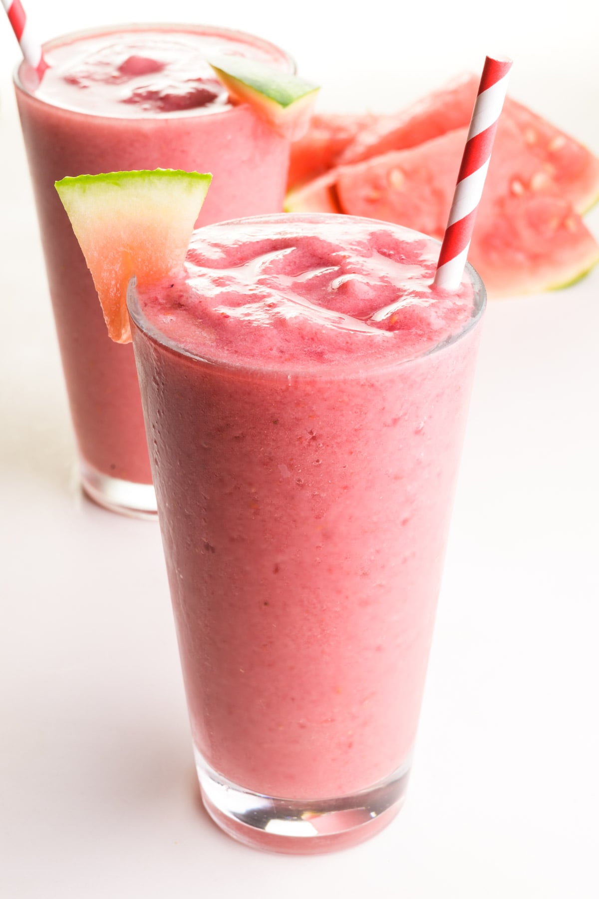 Two pink smoothies sit on a table. They have watermelon wedges and paper straws. There are watermelon wedges in the background.