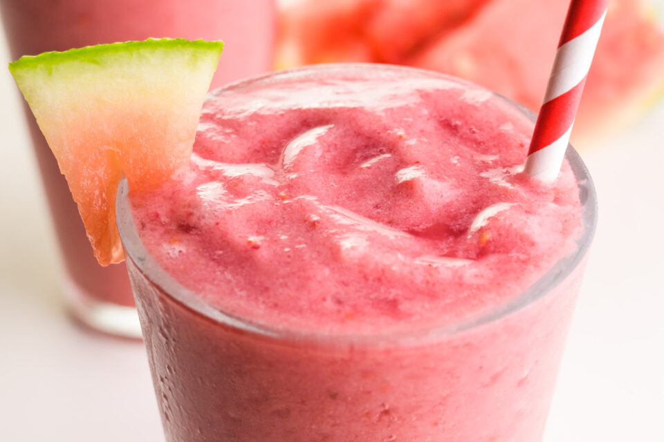 A closeup of a watermelon banana smoothie in a glass. It has a red and white straw and watermelon wedge.