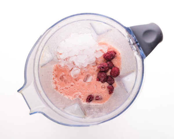 Looking down on a blender jar with pink liquid, raspberries, and crushed ice.