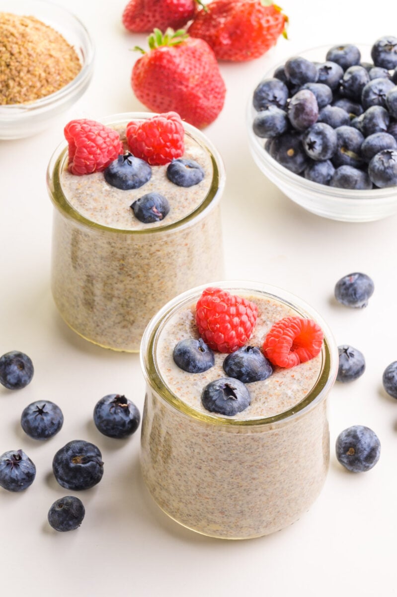 Looking down on two dishes with chia pudding. There are fresh berries on top and around the dishes. There is a bowl of ground flax in the background.