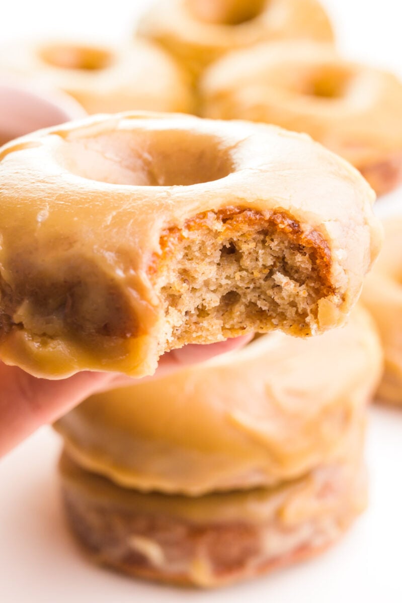 A hand holds a vegan donut with a bite taken out. It hovers over more donuts in the background.