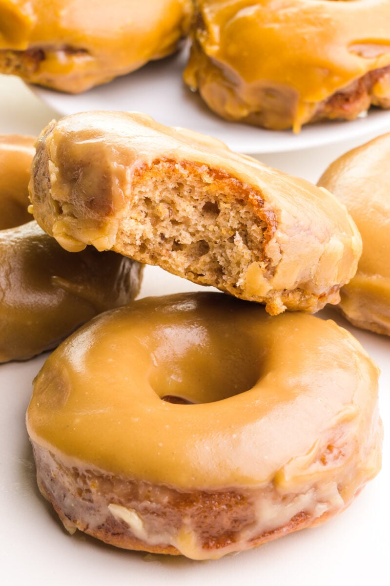 A vegan maple donut with a bite take out sits next to other donuts around it and in the background.