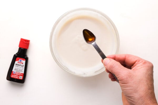 A hand holds a measuring spoon with maple extract over a small bowl with icing in it. There is a bottle of maple extract beside the bowl.